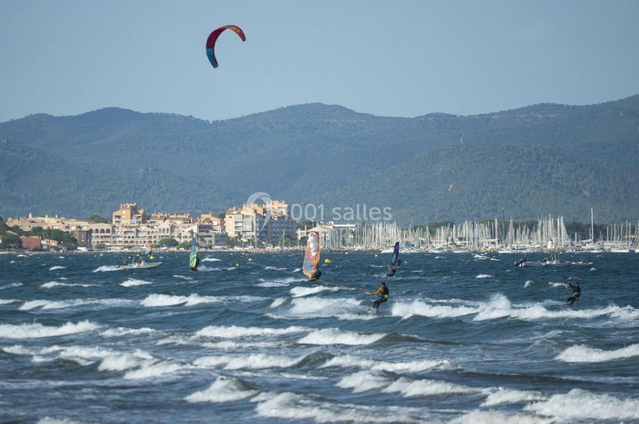 Des personnes pratiquent le kitesurf sur une mer agitée, avec une ville côtière et des collines en arrière-plan.