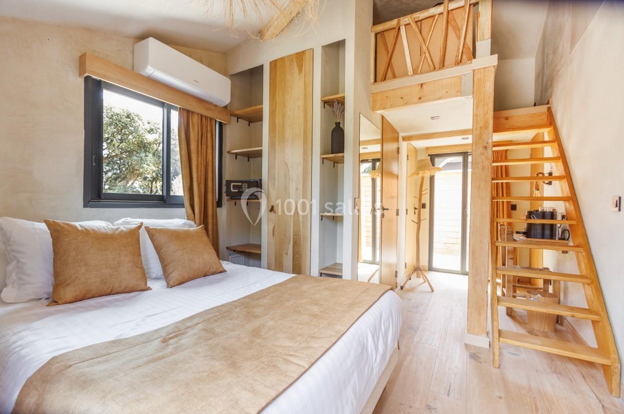 Chambre lumineuse avec lit double, mezzanine en bois, escalier, et grande fenêtre donnant sur un extérieur arboré.