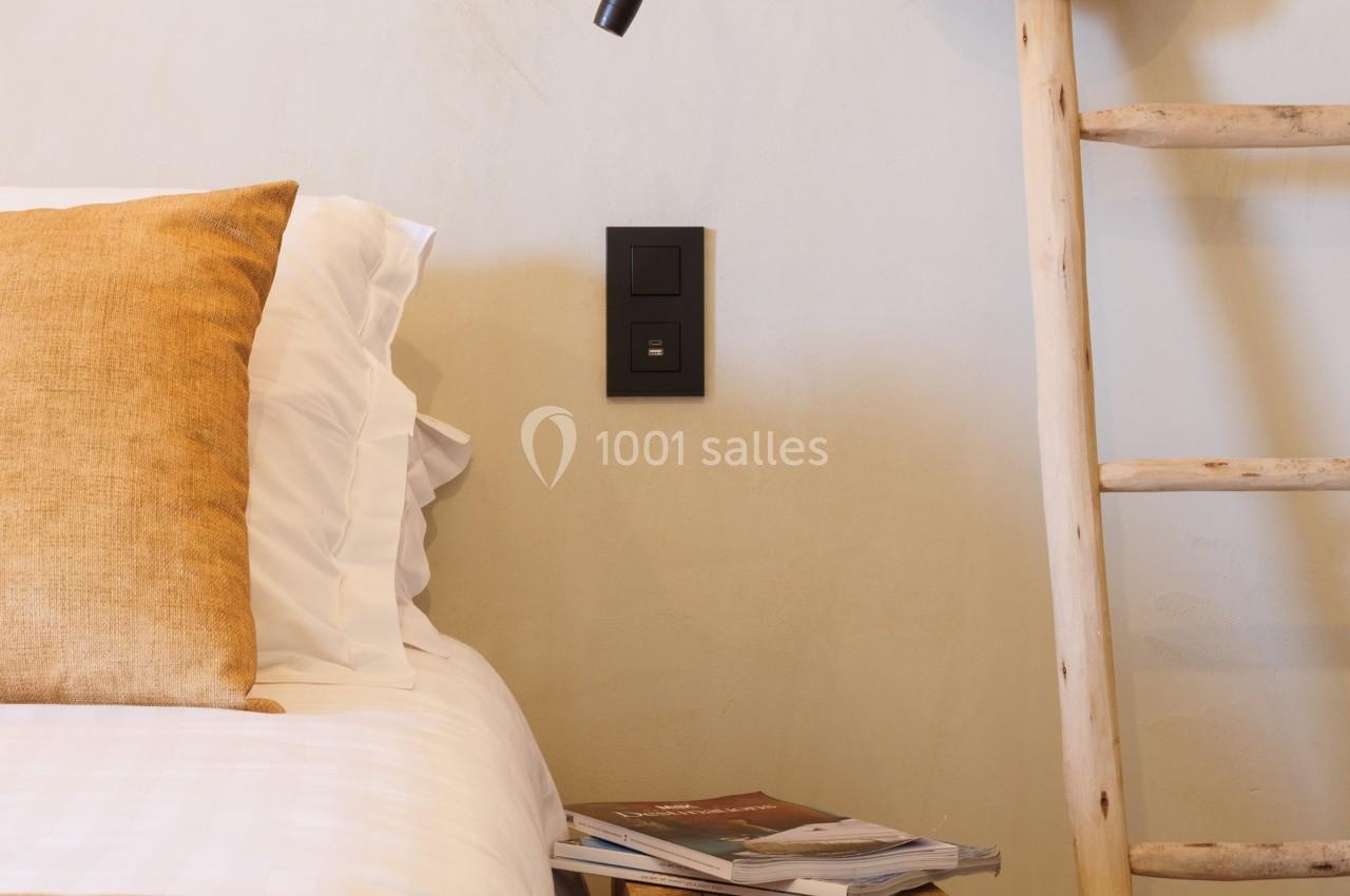 Lit avec oreillers et couverture beige, table de chevet en bois avec magazines, échelle décorative et chapeau en paille.