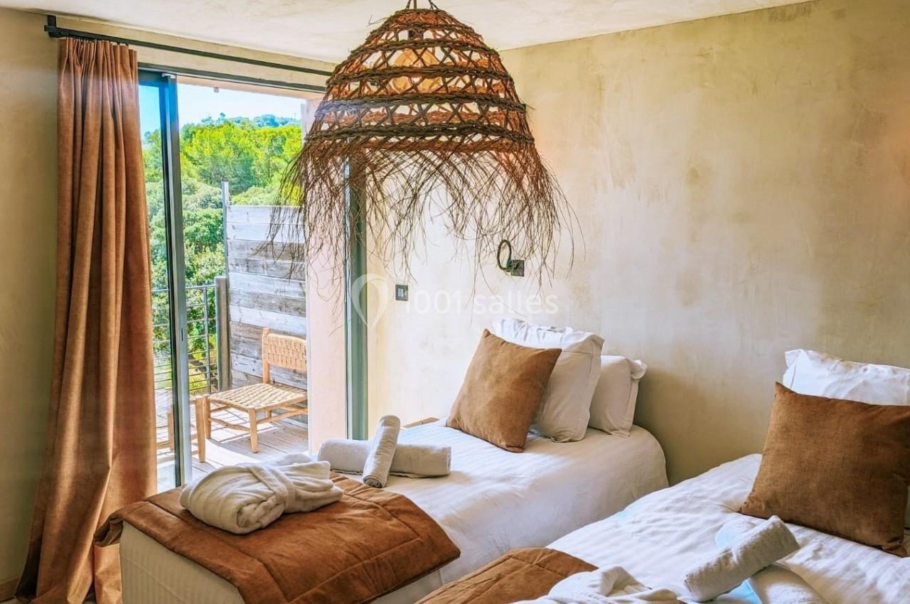 Deux lits simples avec coussins marron dans une chambre lumineuse, ouverte sur une terrasse avec vue sur la végétation.