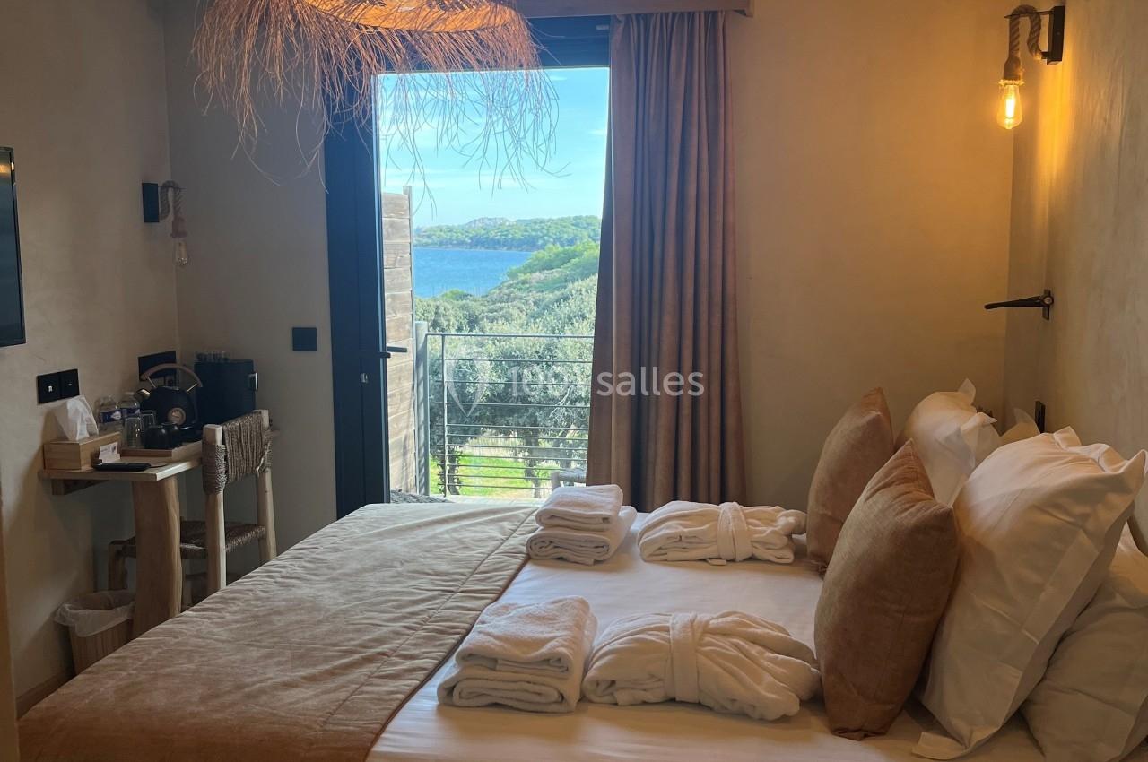 Chambre lumineuse avec lit double, décoration naturelle, vue sur un paysage verdoyant et la mer à travers une porte-fenêtre.