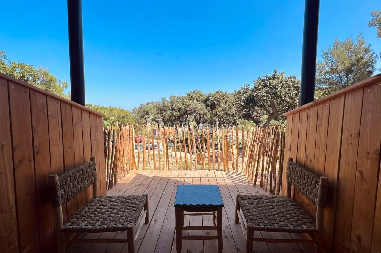 Deux chaises en osier et une table basse sur une terrasse en bois, entourée de palissades et donnant sur un paysage arboré.