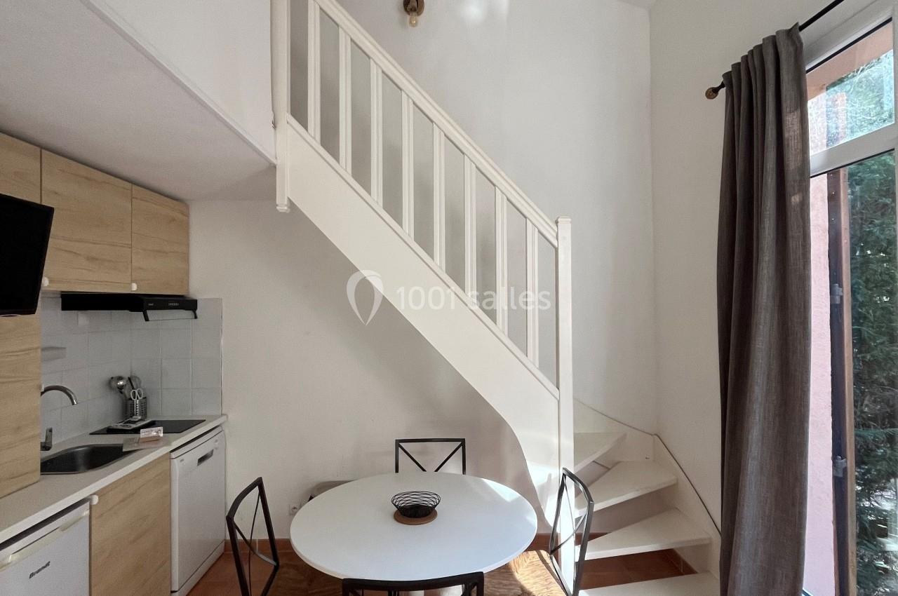 Cuisine ouverte avec table ronde, quatre chaises, escalier blanc, et grande fenêtre avec rideau gris.