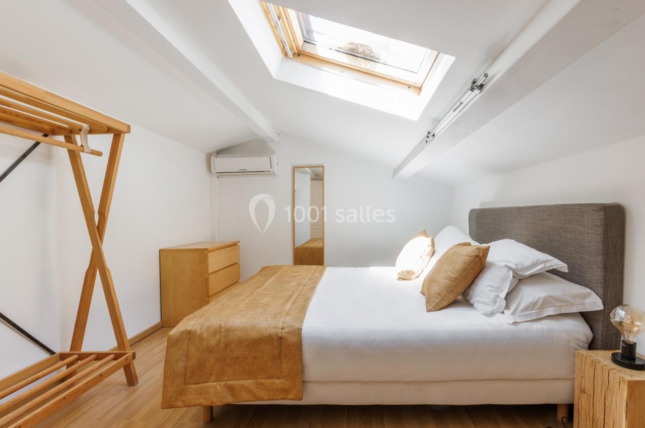 Chambre mansardée lumineuse avec lit double, linge beige, mobilier en bois clair et fenêtre de toit.