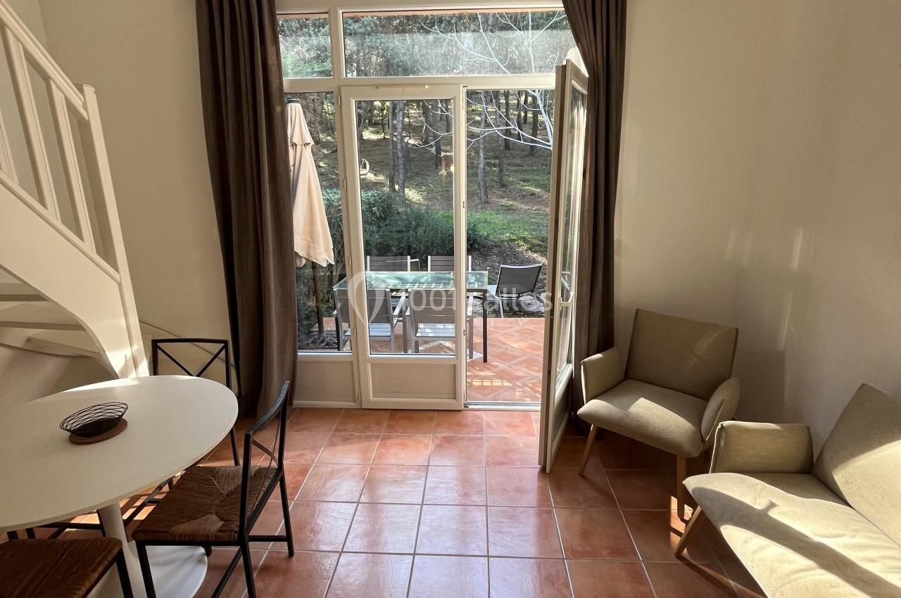 Vue d'un salon lumineux avec carrelage, table ronde, fauteuils, et porte-fenêtre ouverte sur une terrasse et un jardin.