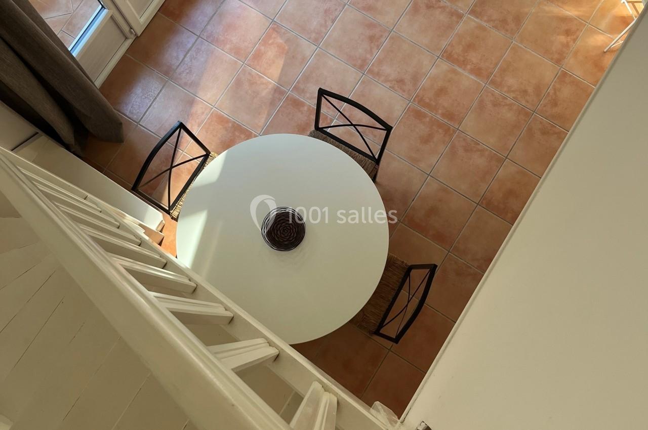 Vue plongeante sur une table ronde blanche entourée de chaises noires, dans un salon avec carrelage et mobilier clair.