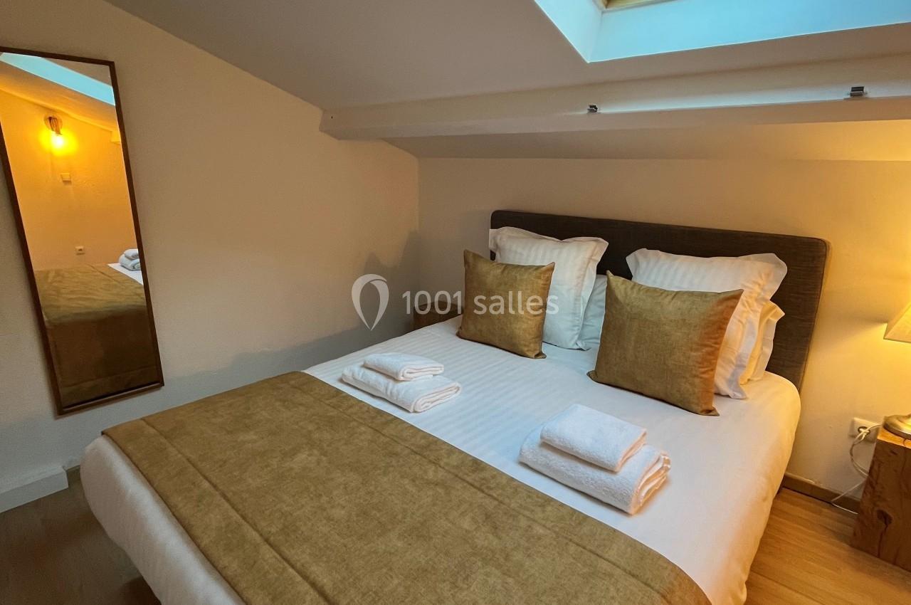 Lit double avec draps blancs et couvre-lit beige, placé sous une fenêtre de toit laissant entrer la lumière naturelle.