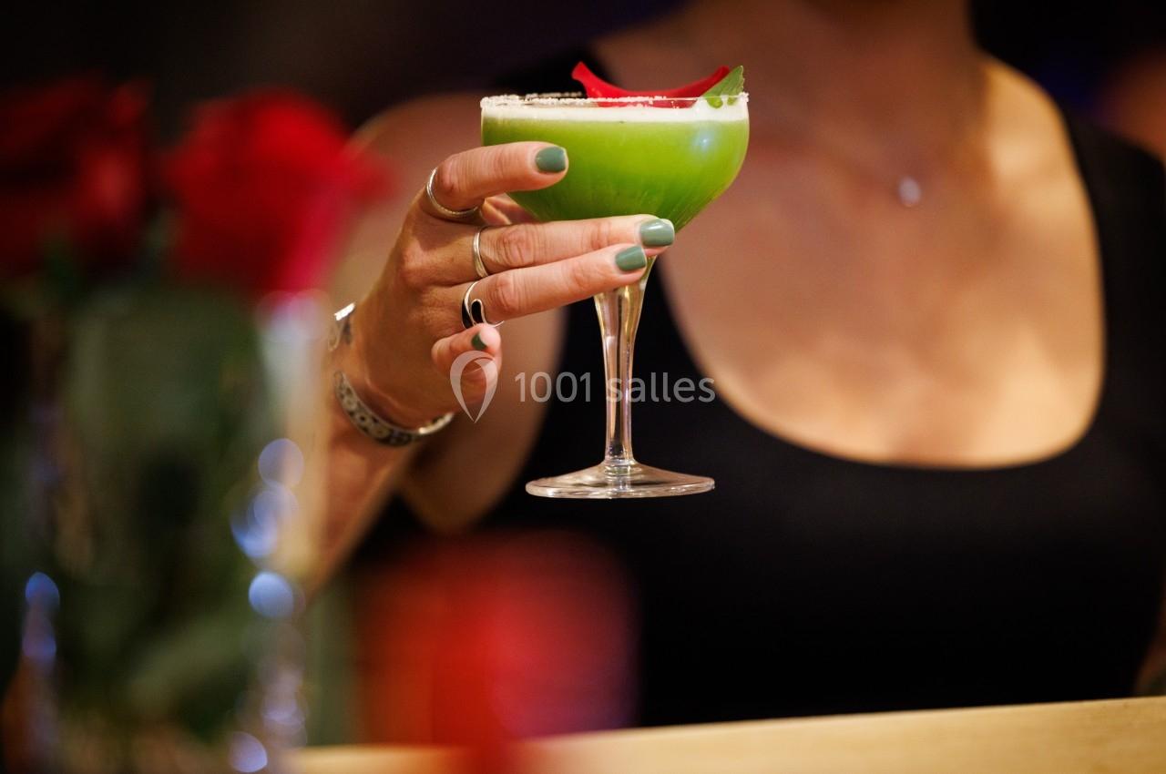 Une personne tenant un cocktail vert garni de pétales rouges dans un verre à pied, en arrière-plan flou.