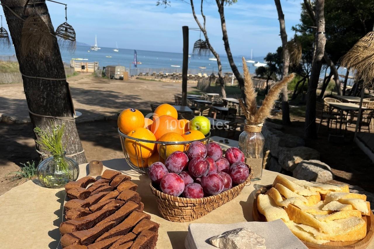 Table avec gâteau au chocolat, fruits frais et pain grillé, en bord de mer sous des pins par temps ensoleillé.