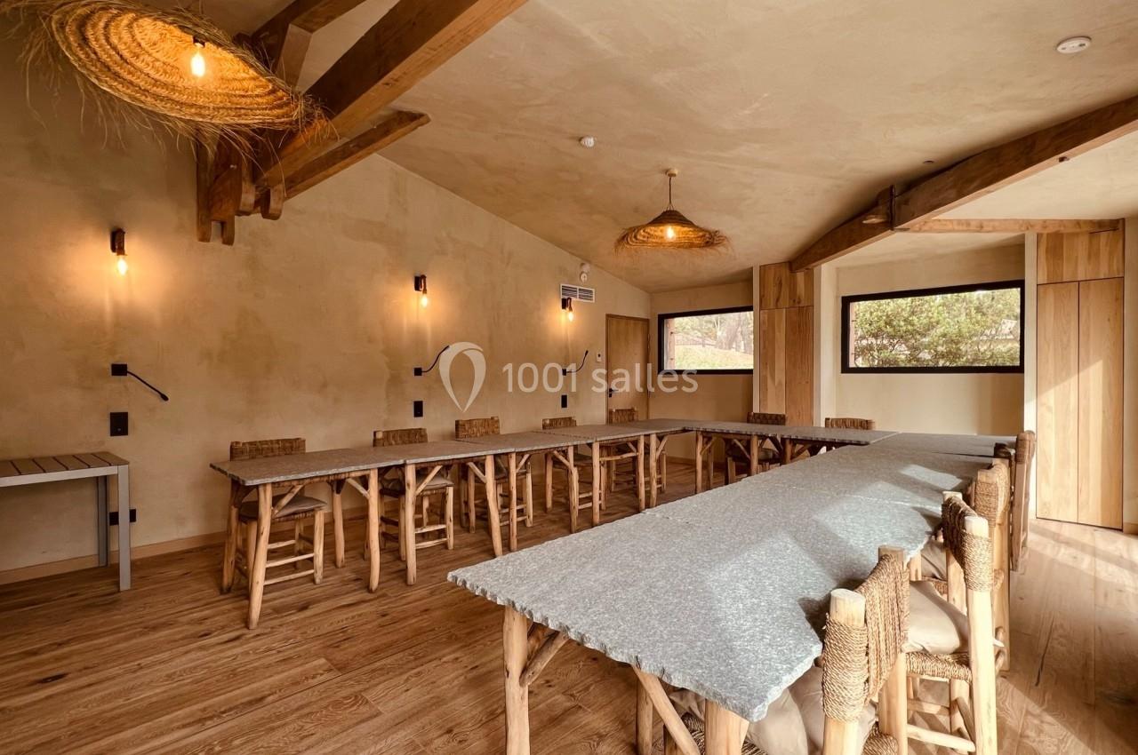 Salle lumineuse avec grandes tables en bois, chaises en osier, poutres apparentes et éclairage suspendu.