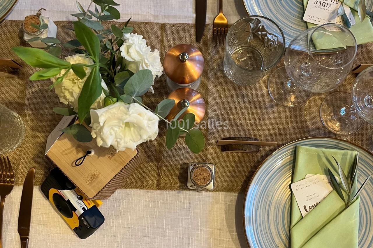Table dressée avec vaisselle en céramique, serviettes vertes, couverts dorés, fleurs blanches et décoration en cuivre.