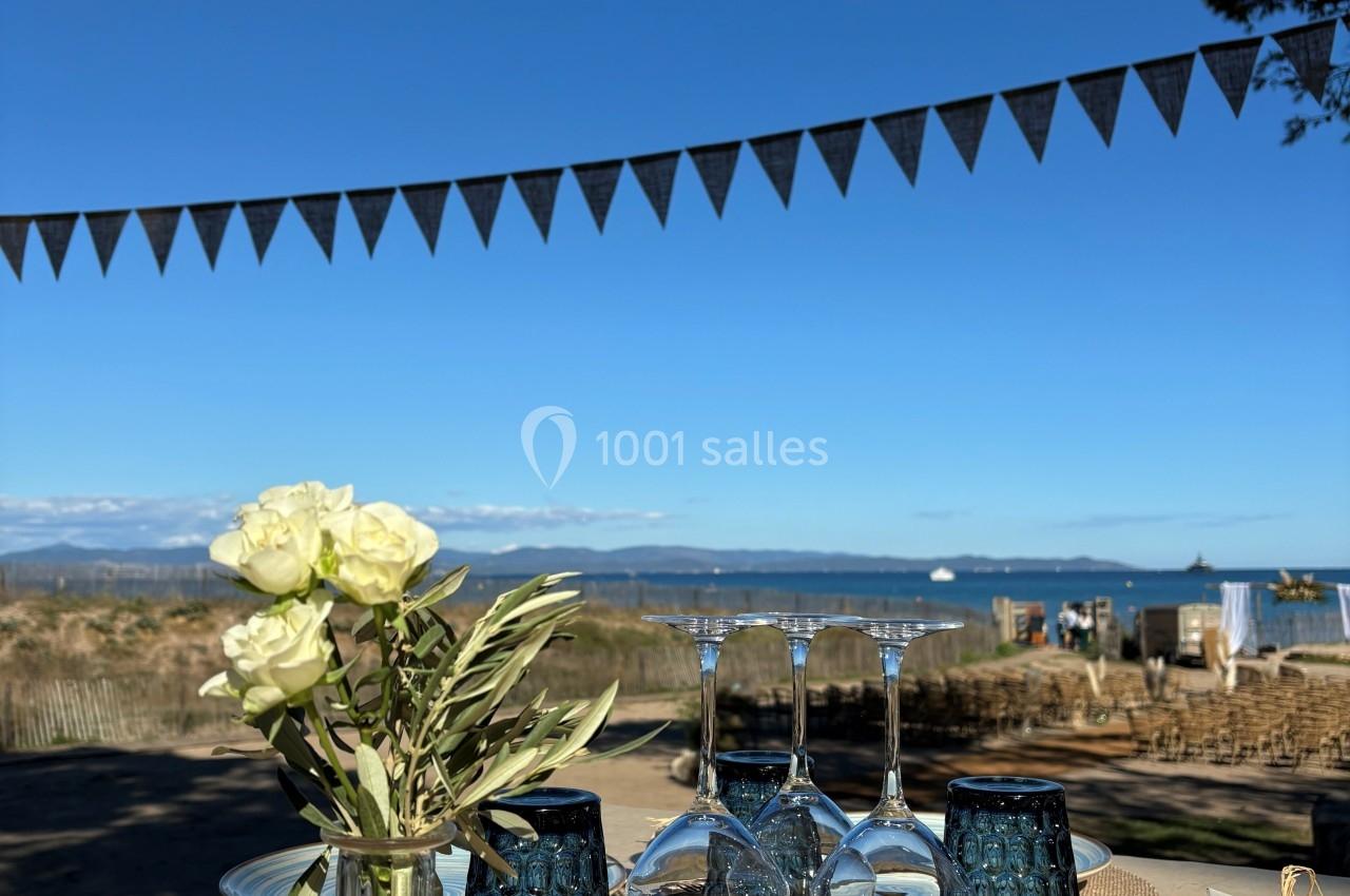 Table extérieure décorée avec des fleurs, vaisselle et verres, avec vue sur la mer et un ciel bleu dégagé.