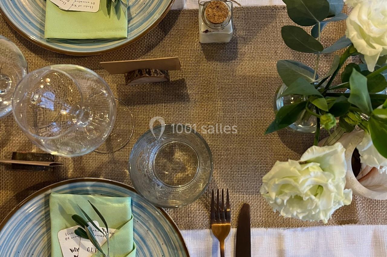 Table dressée avec assiettes en céramique, serviettes vertes pliées, couverts dorés, verres et décoration florale blanche.