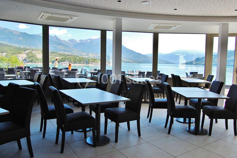 Salle de restaurant avec des tables et chaises noires, offrant une vue panoramique sur un lac et des montagnes.
