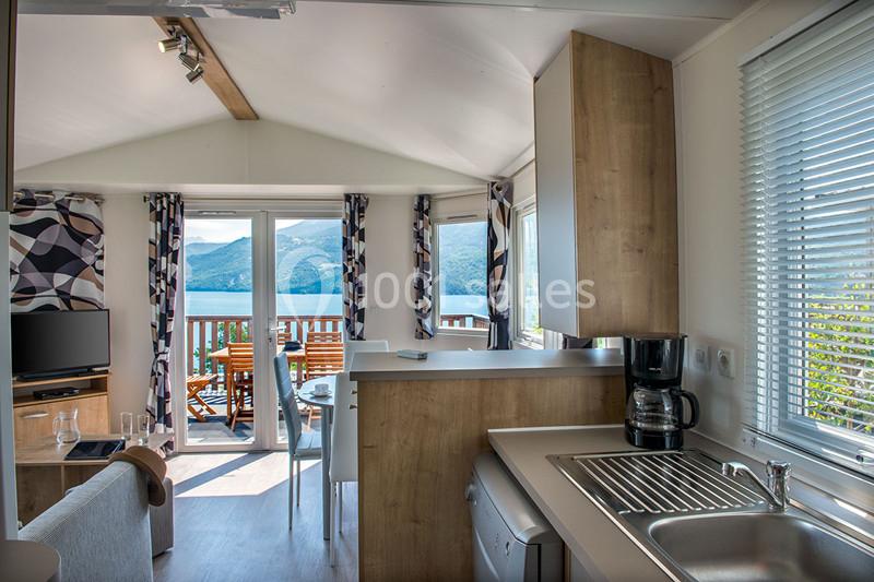 Intérieur d'un mobil-home lumineux avec cuisine équipée, vue sur une terrasse et paysage montagneux à l'extérieur.
