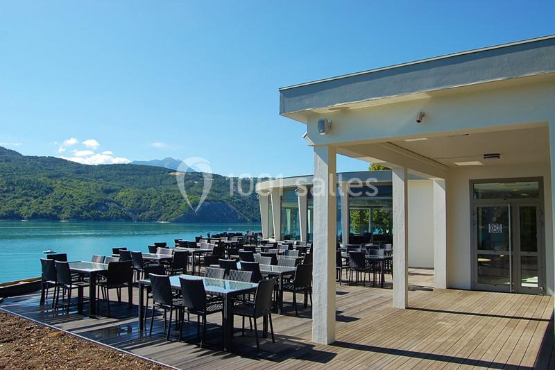 Terrasse d'un restaurant avec tables et chaises, donnant sur un lac entouré de collines sous un ciel dégagé.