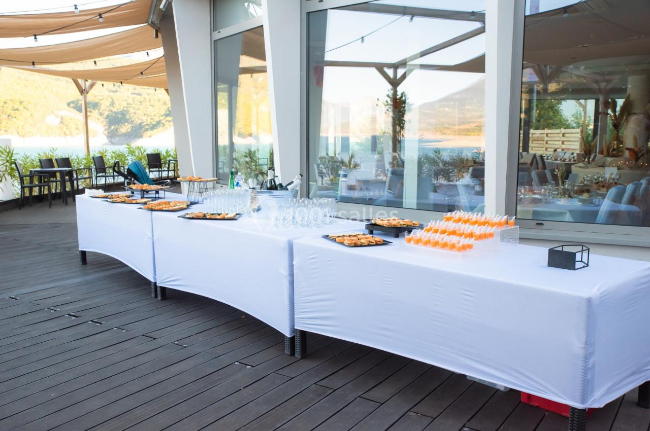 Table de buffet dressée en extérieur avec nappes blanches, verrerie, amuse-bouches et vue sur un paysage naturel.