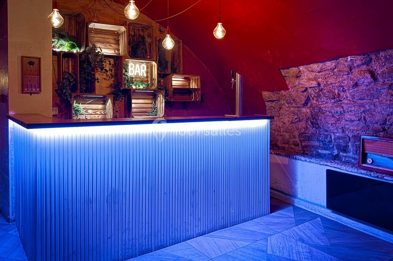 Comptoir de bar rétro éclairé en bleu, avec des étagères décoratives, des ampoules suspendues et un mur en pierre.