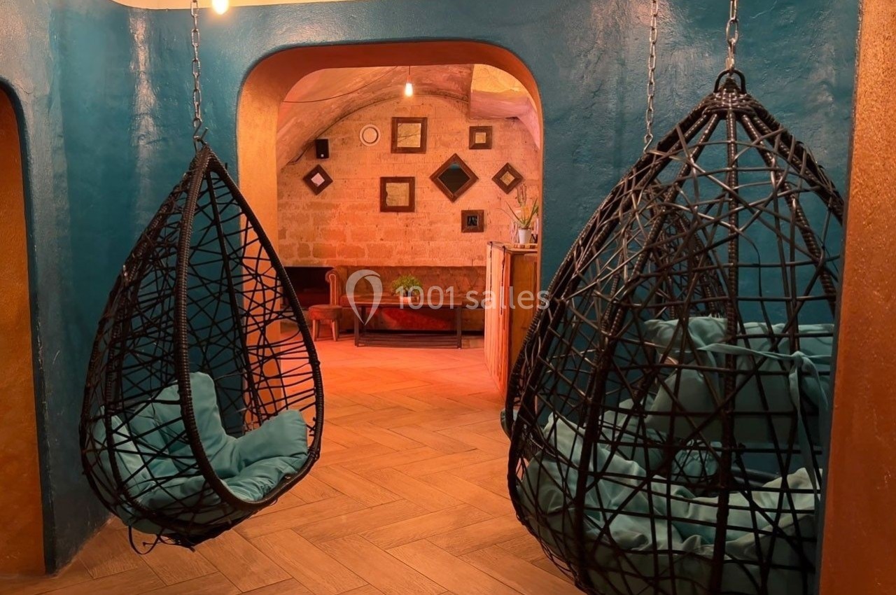 Deux fauteuils suspendus en osier noir avec coussins turquoise dans une pièce aux murs colorés et éclairage chaleureux.