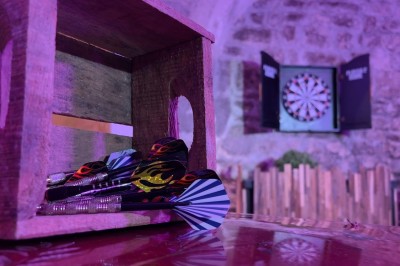 Miniature Location salle Paris 4 (Paris) - Paris Clandestin #31 Fléchettes colorées dans une caisse en bois au premier plan, cible de jeu accrochée sur un mur en pierre en arrière-plan.