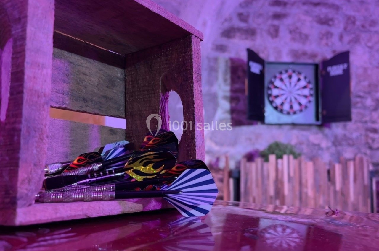 Fléchettes colorées dans une caisse en bois au premier plan, cible de jeu accrochée sur un mur en pierre en arrière-plan.