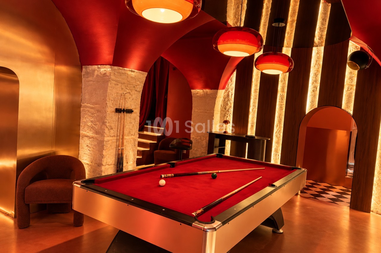 Salle de billard au plafond voûté rouge, avec table à tapis rouge, plusieurs suspensions colorées, murs dorés et parois en pierre éclairées.