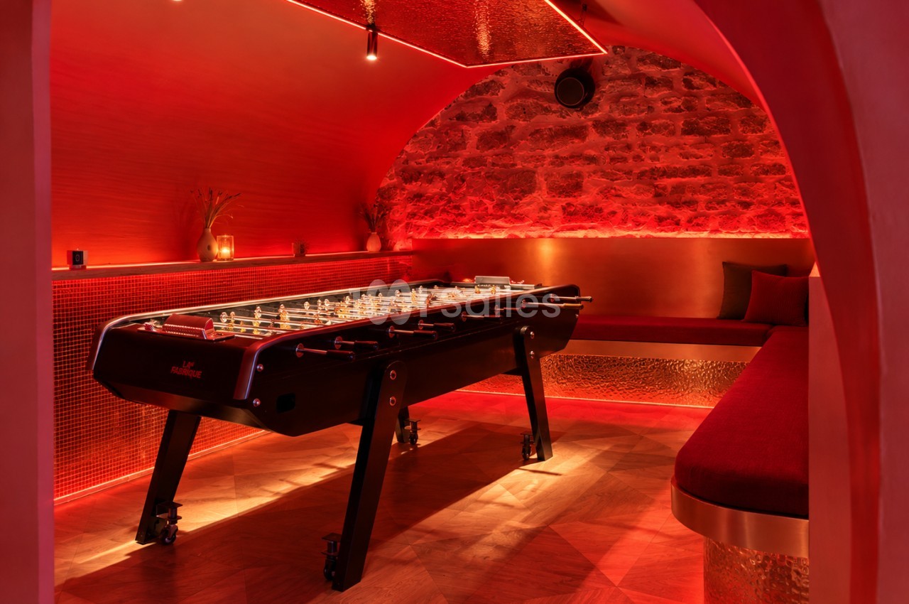 Baby-foot noir installé dans une salle voûtée aux murs de pierre et de carrelage mosaïque, éclairée en rouge, avec banquettes rouges en arrière-plan.