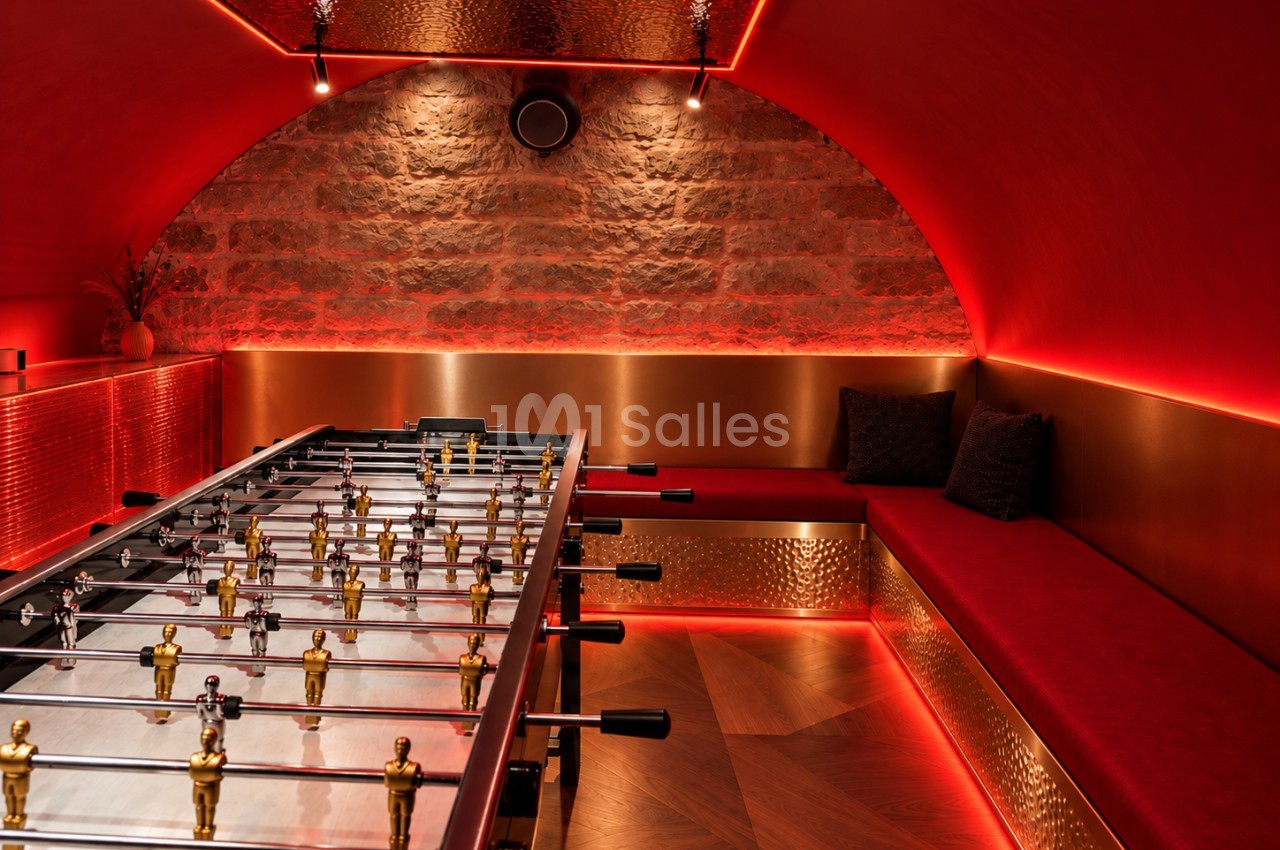 Salle voûtée aux murs rouges éclairés, avec un baby-foot doré au premier plan et une banquette rouge le long d'un mur de pierre apparente.
