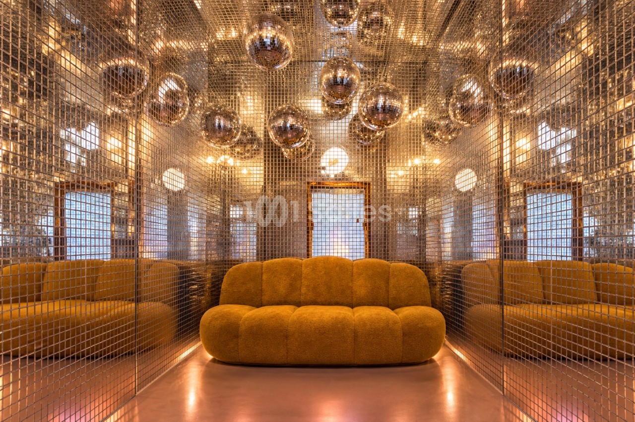 Salon aux parois en grille métallique dorée reflétant des boules à facettes suspendues, avec un canapé capitonné couleur moutarde au centre.