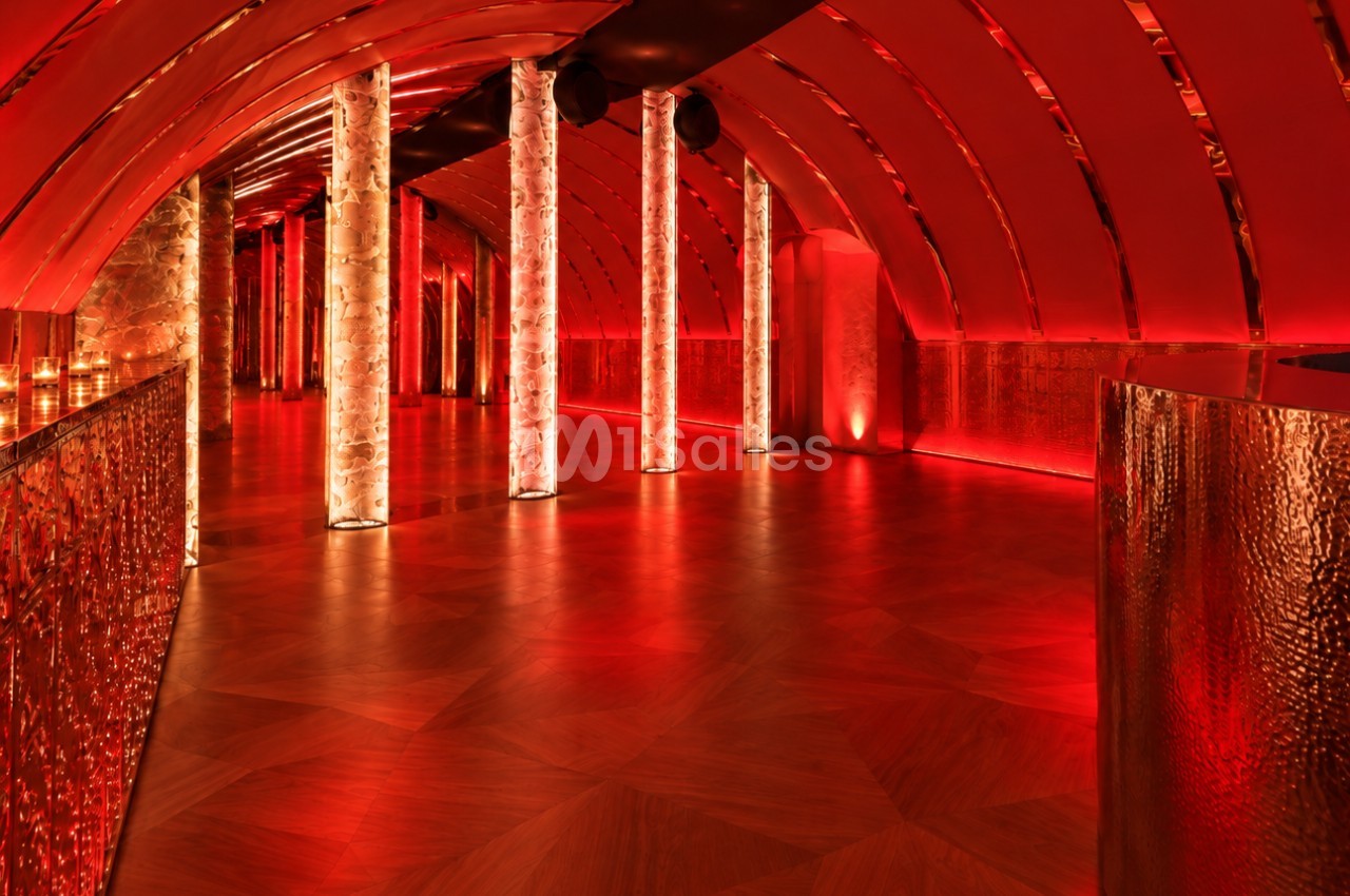 Salle voûtée à l'éclairage rouge avec colonnes lumineuses blanches, sol en parquet et longue allée centrale sans mobilier visible.