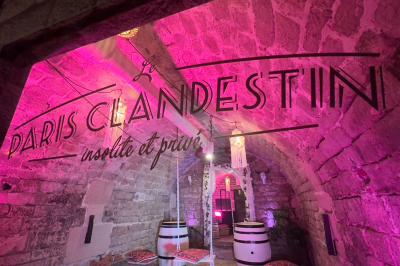 Miniature Le Fumoir Fléchettes colorées dans une caisse en bois au premier plan, cible de jeu accrochée sur un mur en pierre en arrière-plan.