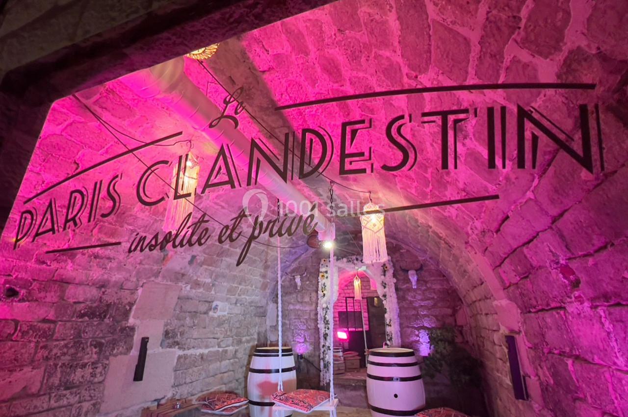 Salle voûtée en pierre éclairée de lumières roses, avec des tonneaux et une inscription ’Paris Clandestin’.