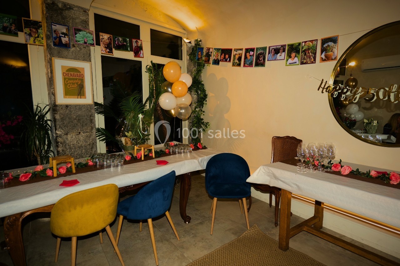 Salle décorée pour une fête avec guirlandes de photos, ballons, fleurs et tables dressées avec nappes blanches.