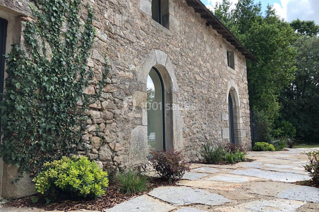 Façade en pierre d'une maison ancienne avec arches, végétation et allée pavée dans un cadre naturel.