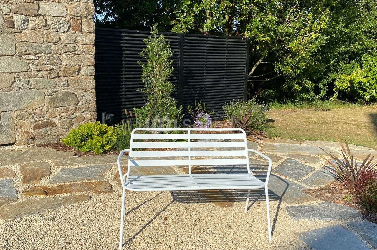 Banc en métal blanc placé sur une terrasse en pierre, entouré de végétation et d'un mur en pierre.