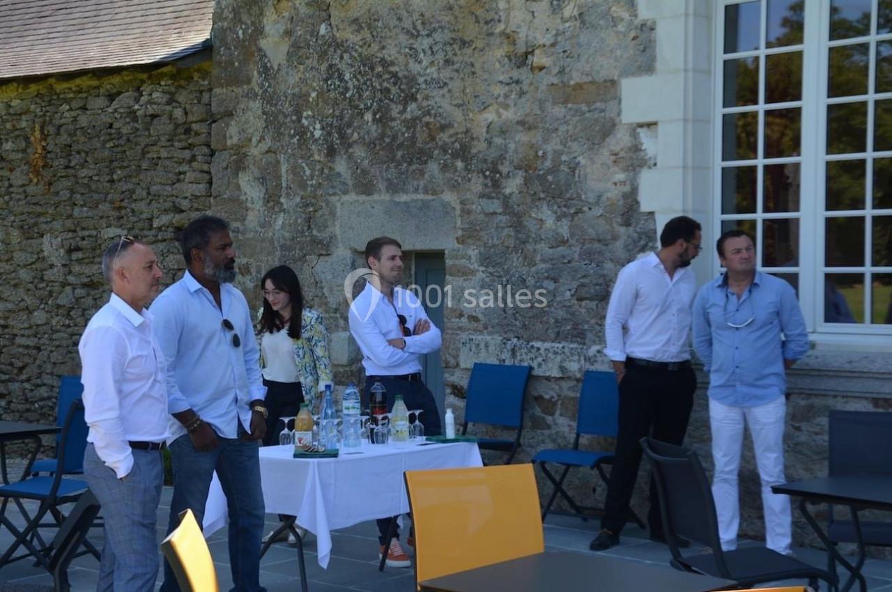 Un groupe de personnes debout en extérieur près d'une table avec des boissons, devant un mur en pierre et une grande fenêtre.