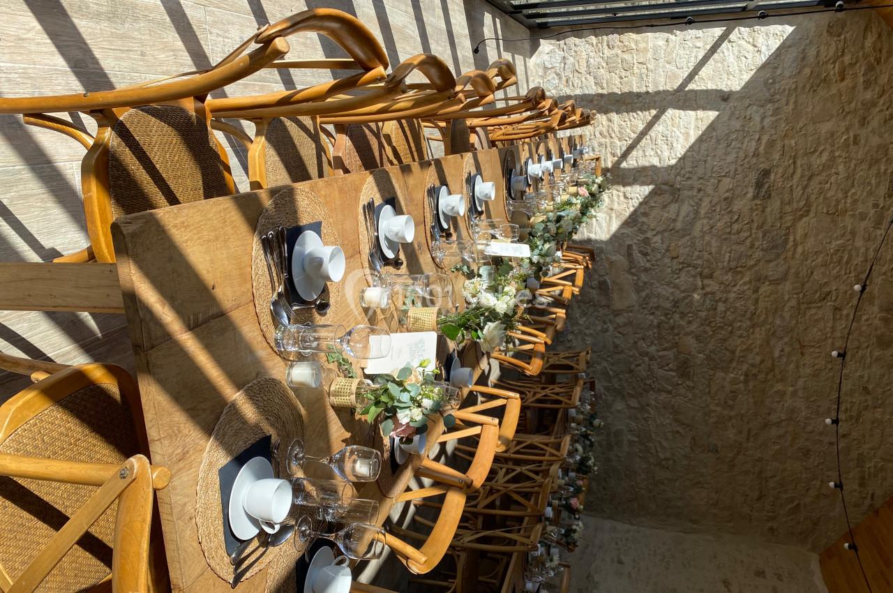 Table en bois dressée pour un repas, avec vaisselle blanche, décor floral et lumière naturelle entrant par une verrière.