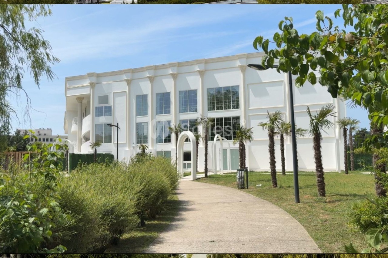 Bâtiment blanc moderne entouré de verdure, avec un chemin piéton bordé d'arbustes menant à l'entrée principale.