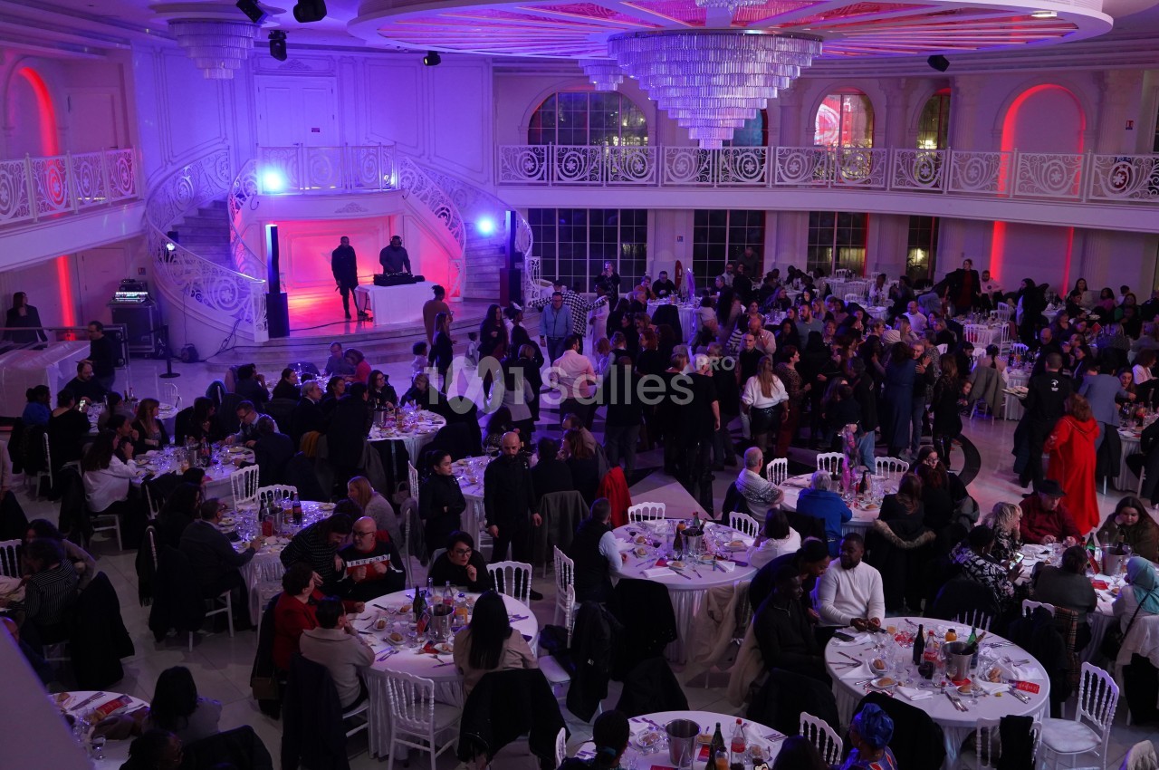 Salle de réception éclairée en violet et rouge, avec des tables rondes occupées par de nombreux convives.