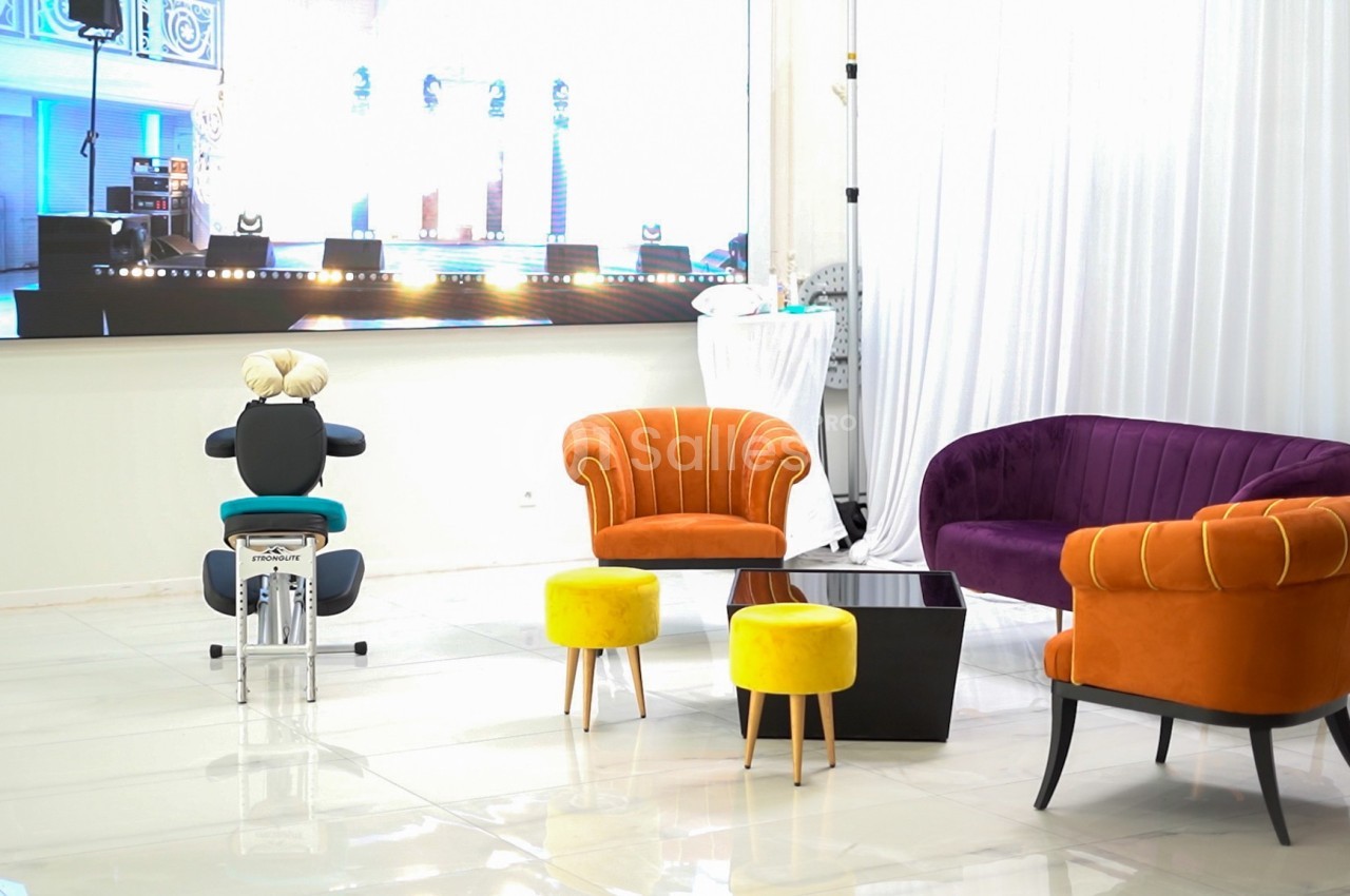 Salle lumineuse avec fauteuils colorés, tabourets jaunes, table basse noire et écran lumineux en arrière-plan.