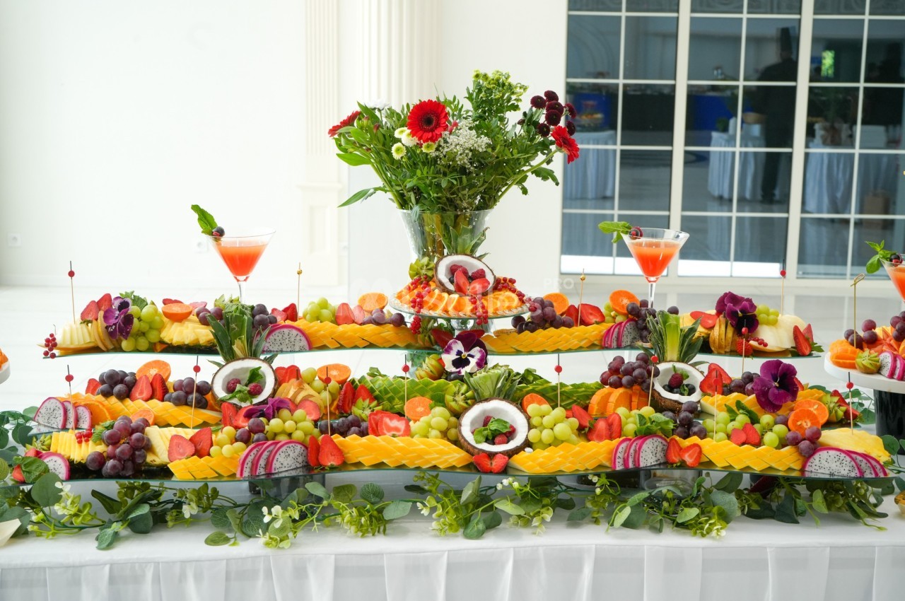 Plateau de fruits variés joliment disposés sur une table, avec des décorations florales et des cocktails en arrière-plan.