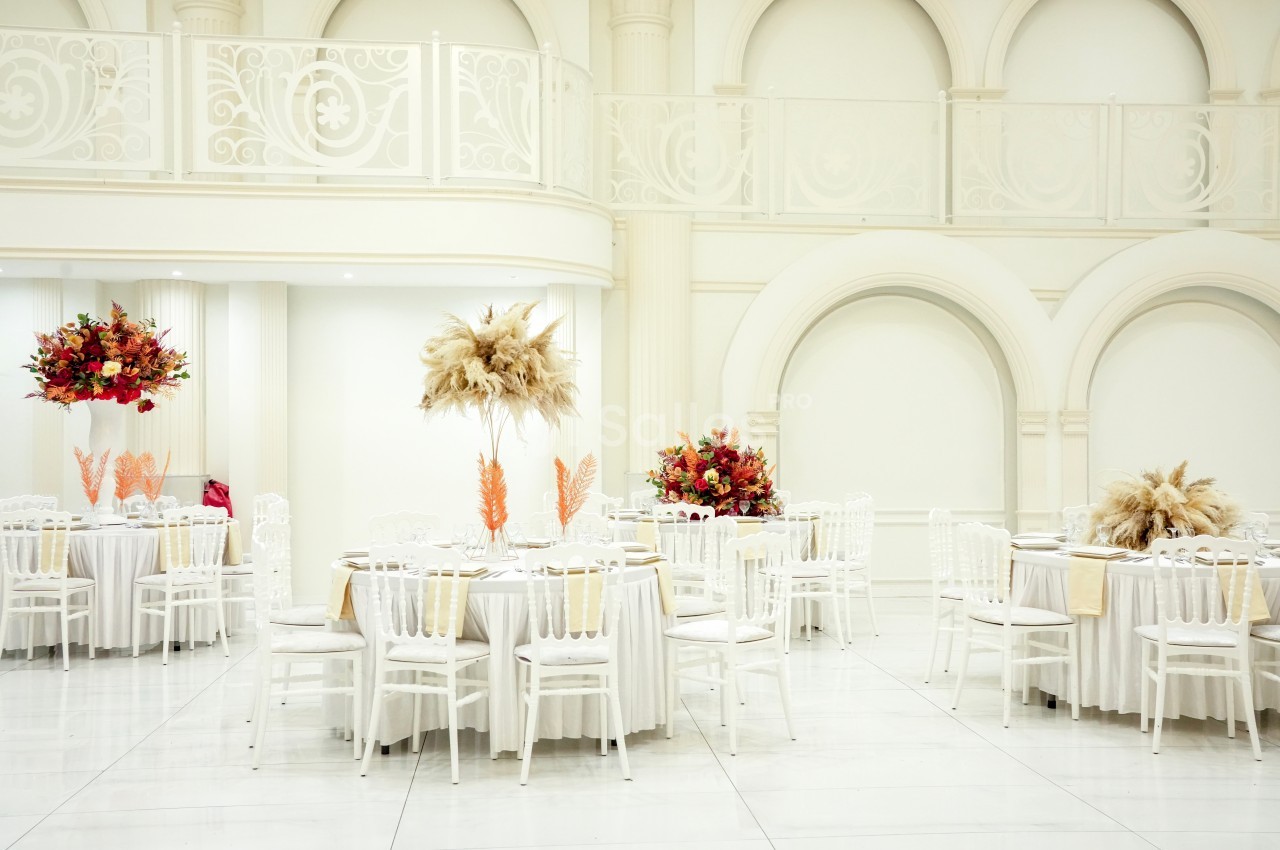 Salle de réception lumineuse avec tables rondes décorées de fleurs colorées et de pampas, entourées de chaises blanches.
