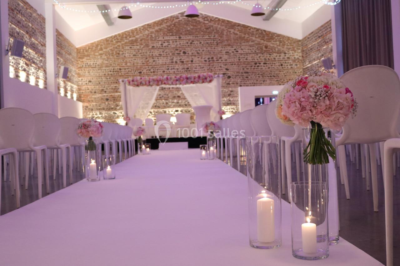 Allée centrale décorée de fleurs et bougies menant à une arche ornée de fleurs dans une salle en briques apparentes.