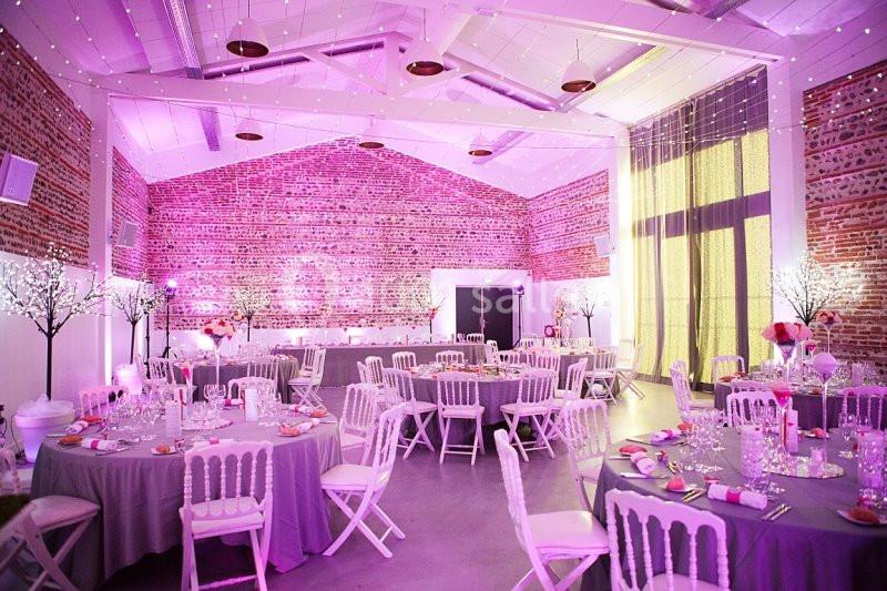 Salle de réception décorée avec des tables rondes, nappes grises, chaises blanches et éclairage rose tamisé.
