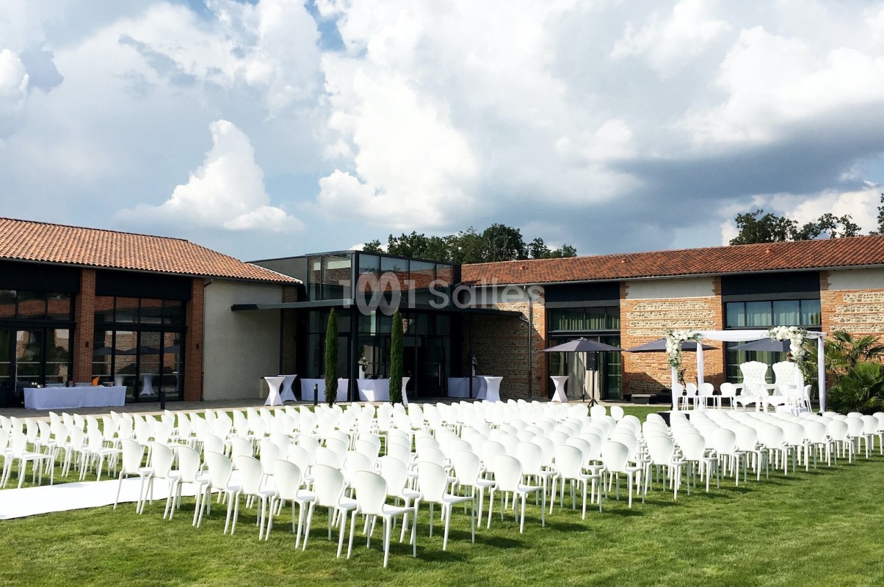 Chaises blanches alignées en extérieur devant un bâtiment moderne en briques, sous un ciel partiellement nuageux.