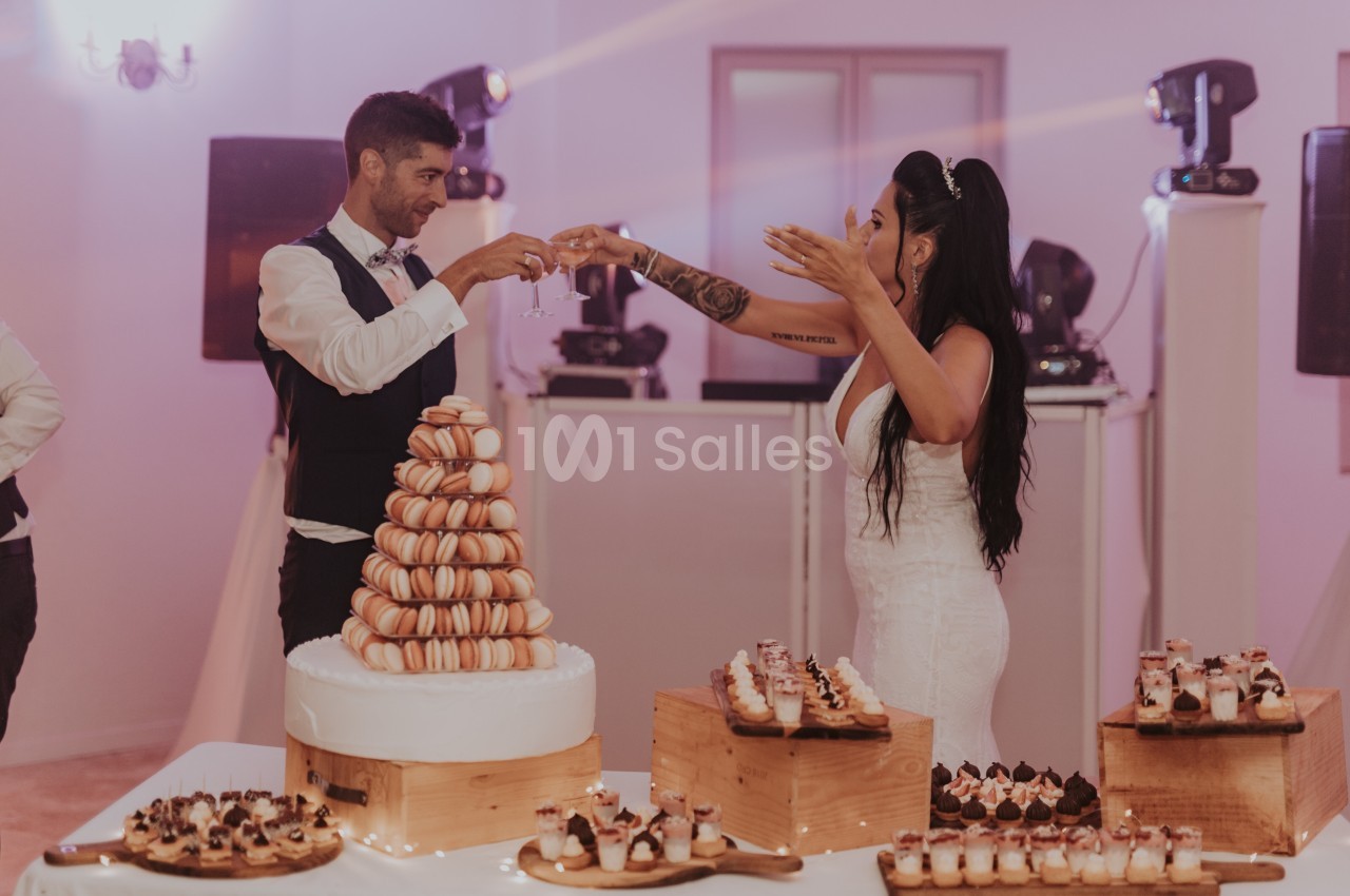 Un couple en tenue de mariage trinque devant une pièce montée de macarons et des desserts sur des tables décorées.