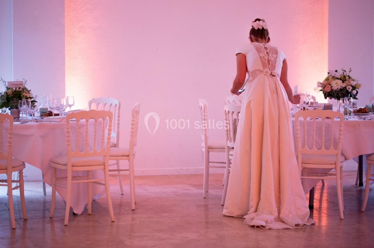 Une femme en robe de mariée ajuste une table décorée pour un événement dans une salle éclairée en rose.
