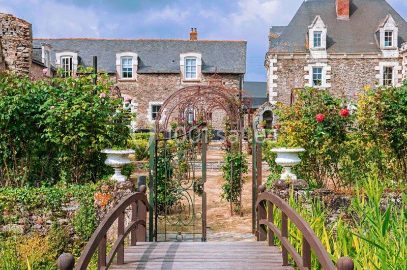 Jardin fleuri avec une arche métallique, un pont en bois et des bâtiments en pierre en arrière-plan.