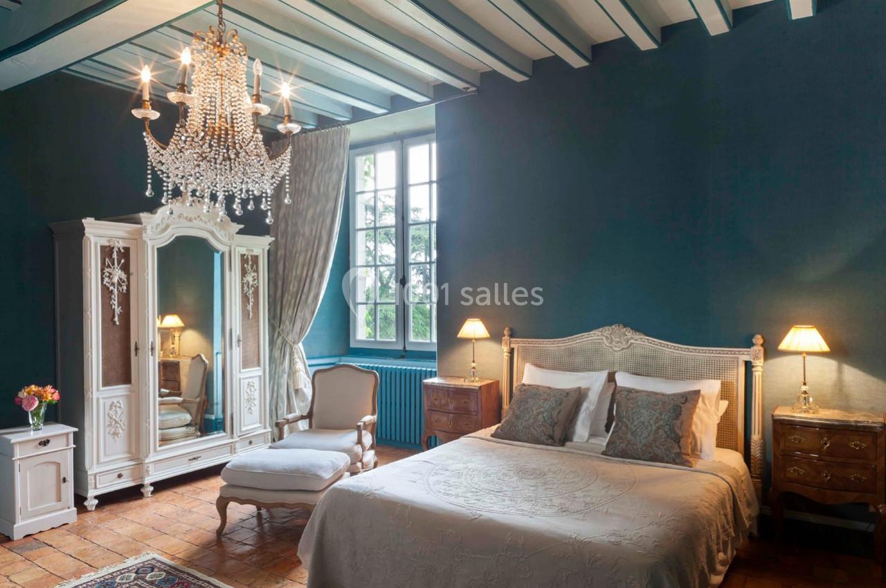 Chambre élégante avec lit double, armoire ancienne, fauteuil, lustre en cristal et mur bleu, éclairée par une grande fenêtre.