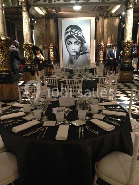 Salle élégante avec tables rondes dressées, chaises blanches et grand portrait en noir et blanc en arrière-plan.